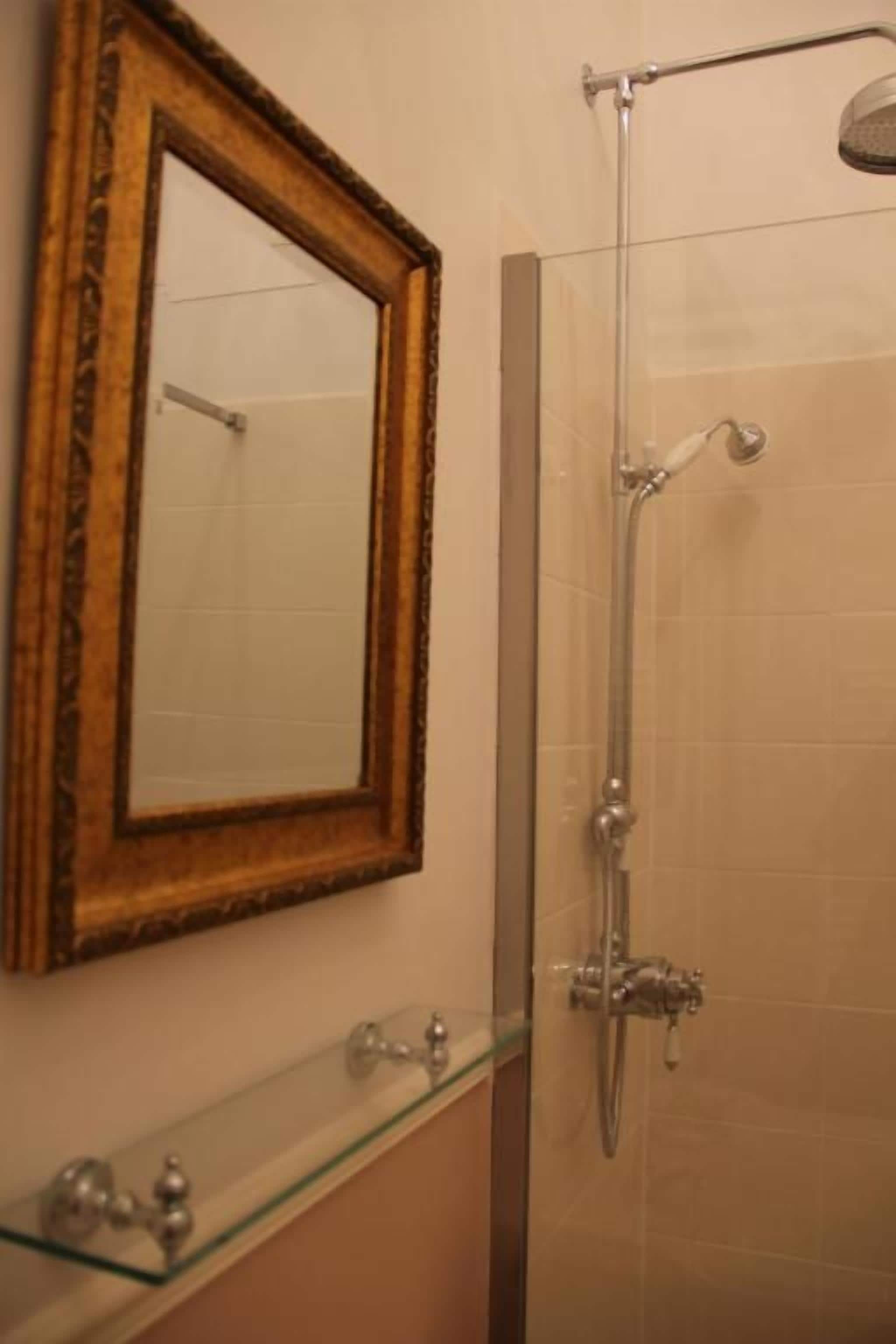 A bathroom with a glass shower enclosure, a wooden-framed mirror, and a glass shelf with a faucet.