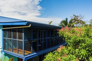 Eine blaue, glasüberdachte Konstruktion mit einer Holzterrasse, umgeben von üppiger Vegetation und Palmen.