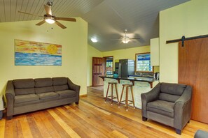 Una sala de estar con piso de madera, un sofá marrón, una silla de madera, una zona de cocina con refrigerador y un ventilador de techo.