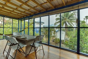 Un espacio al aire libre cubierto con comedor, mesa, sillas, ventanas amplias y vistas a una casa y palmeras.