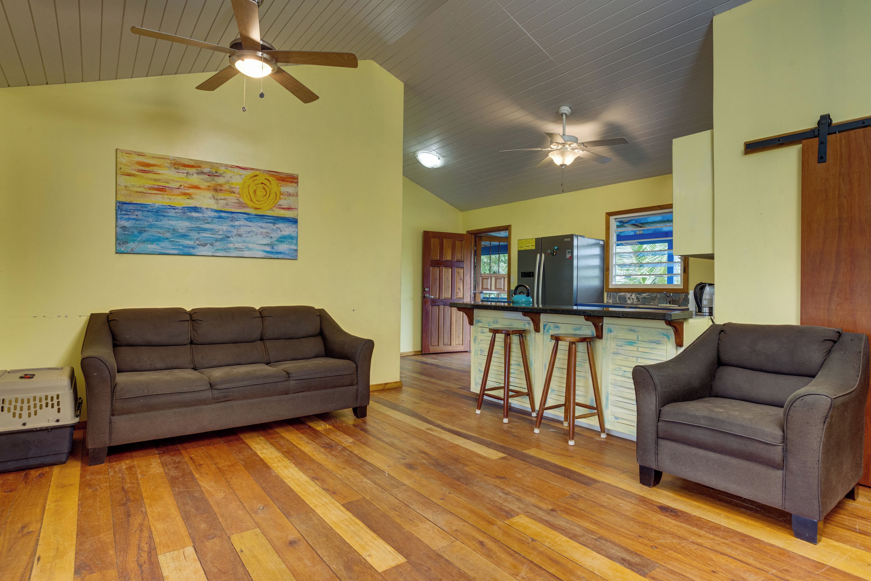 A spacious living area with a wooden floor, a sofa, a chair, and a kitchen with a refrigerator and bar stools.