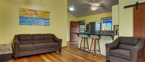 A spacious living area with a wooden floor, a sofa, a chair, and a kitchen with a refrigerator and bar stools.