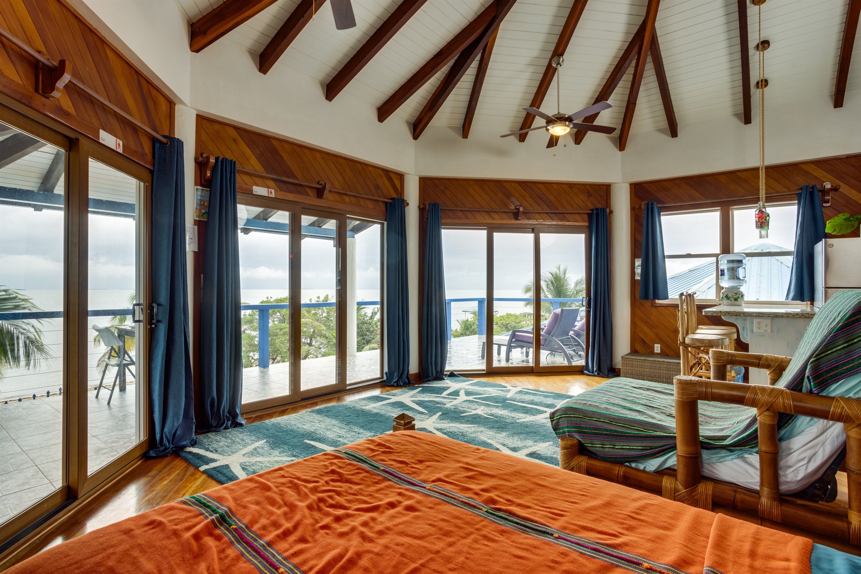 A spacious room with a large sliding glass door leading to a balcony, a bed with a colorful blanket, a chair, and a ceiling fan.