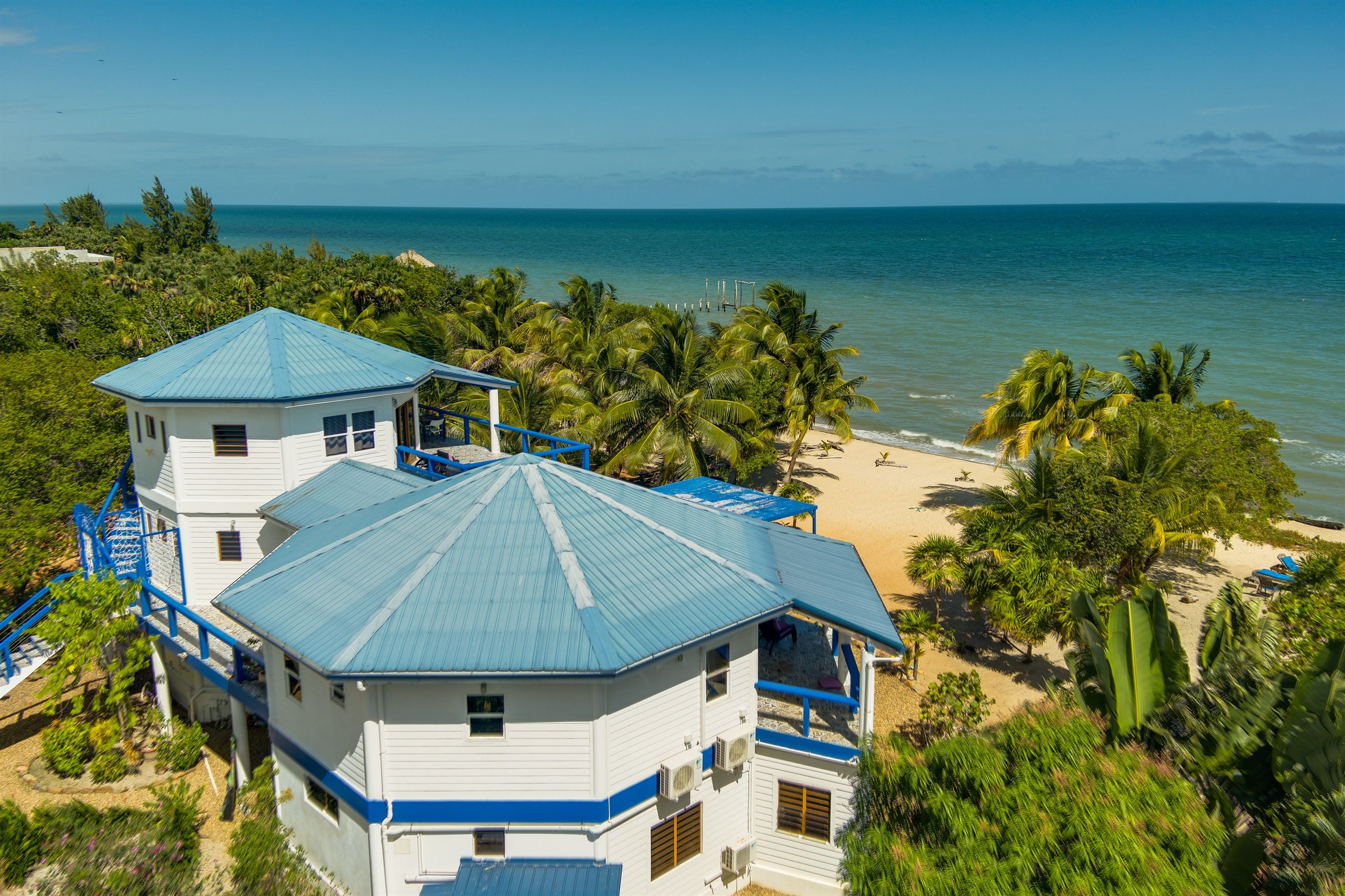 A coastal property with a large building featuring multiple levels and a prominent blue roof, surrounded by lush greenery and a beach.