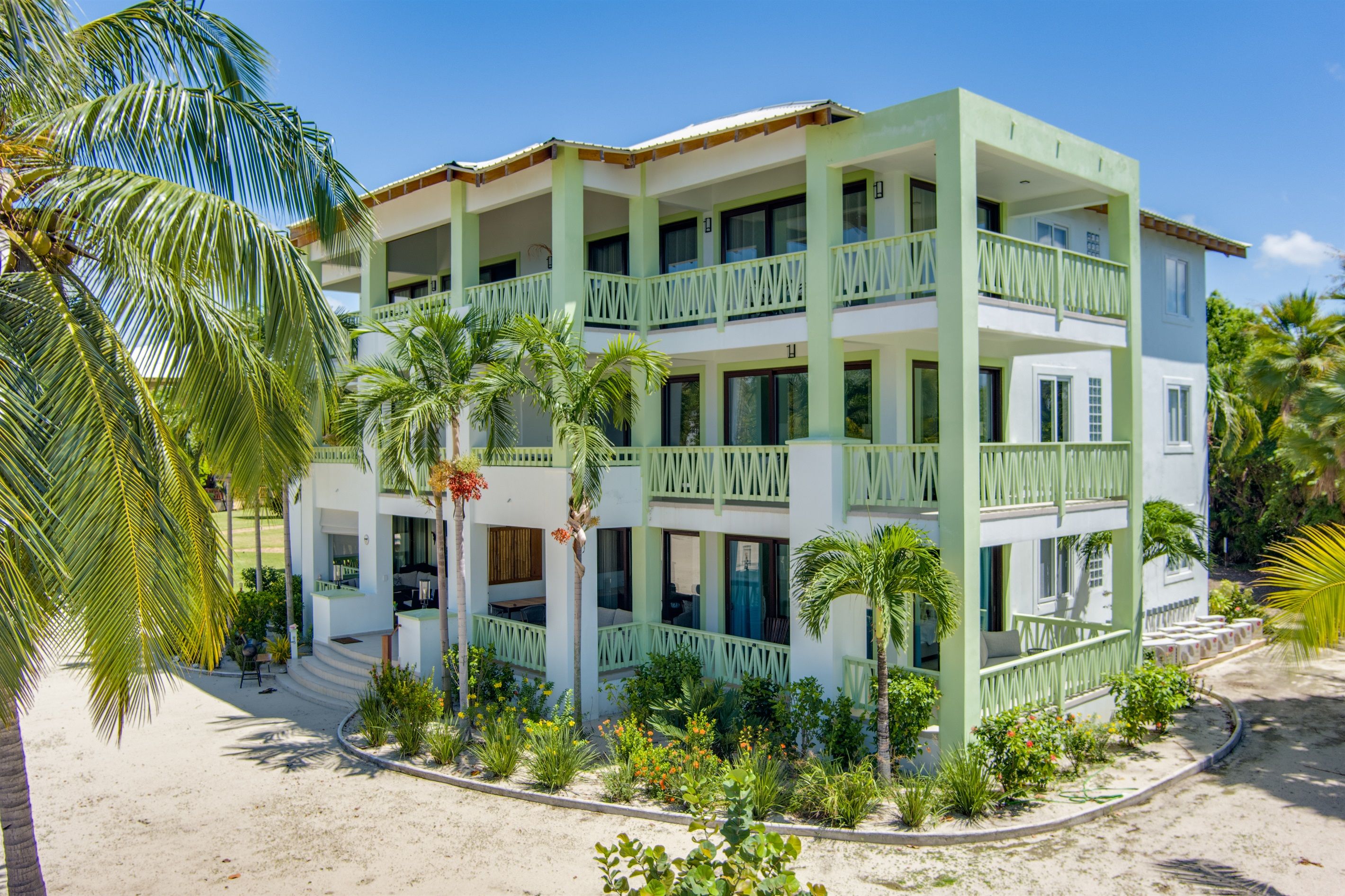 A multi-story hotel with a green and white color scheme, featuring balconies and a landscaped garden with palm trees.