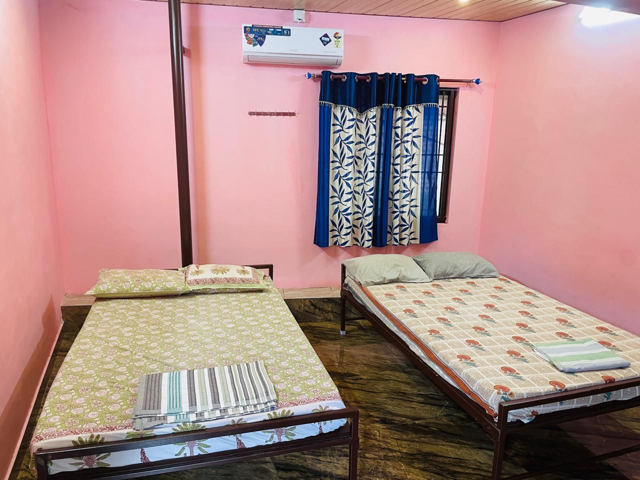 Una habitación con dos camas individuales, una ventana con cortinas azules, un equipo de aire acondicionado y un estante fijado a la pared.