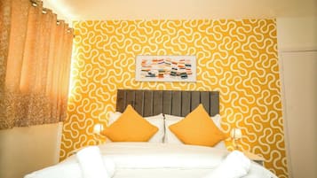 Une chambre d’hôtel avec un mur jaune à motifs, un lit avec une literie blanche et deux oreillers orange, et une œuvre d’art encadrée au-dessus du lit.