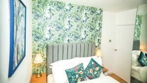 Ein Schlafzimmer mit einem Bett, Kissen, einem Nachttisch, einer Lampe, einer Wand mit tropischem Blattmuster und einer Tür.