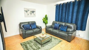 Ein Wohnzimmer mit zwei Ledersofas, einem Glastisch, einem grünen Teppich, einer Weltkarte als Kunstwerk und blauen Vorhängen.