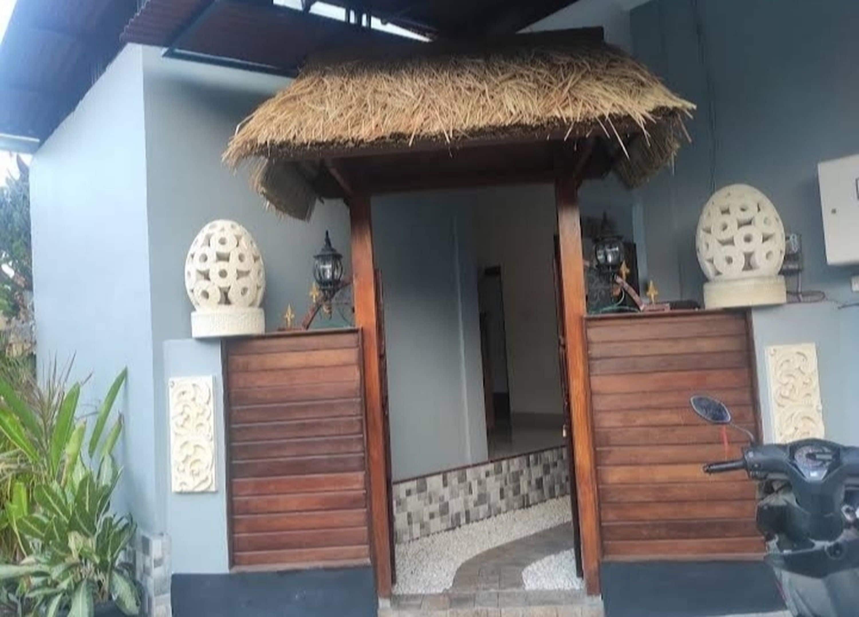 A house entrance with a thatched roof overhang, wooden doors, decorative spheres, and a scooter parked outside.