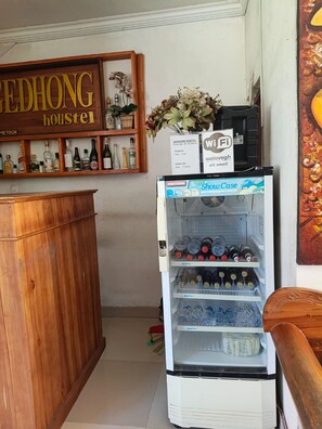 Une chambre d’hôtel avec un comptoir d’accueil en bois, un réfrigérateur rempli de boissons, une enseigne indiquant « Gedhong Hostel » et un symbole Wi-Fi.