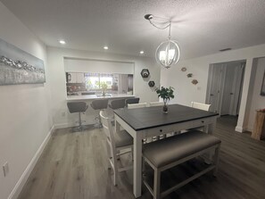 A modern dining area with a dark wooden table, white chairs, and a bench. There is a chandelier above the table and a potted plant on the table. - Cozy 2-bedroom condo in peaceful Port Richey with AC (Port Richey)