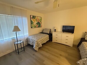 A bedroom with a bed, a bedside table with a lamp, a television, and a painting on the wall. - Cozy 2-bedroom condo in peaceful Port Richey with AC (Port Richey)
