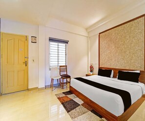 A hotel room with a large bed, a wooden door, a window with blinds, a chair, and a small table with a vase of flowers. - Classic Goldens Residency (Puducherry)