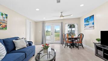 A living room with a blue sofa, a glass coffee table, a dining area with a table and chairs, a ceiling fan, and a TV stand.
