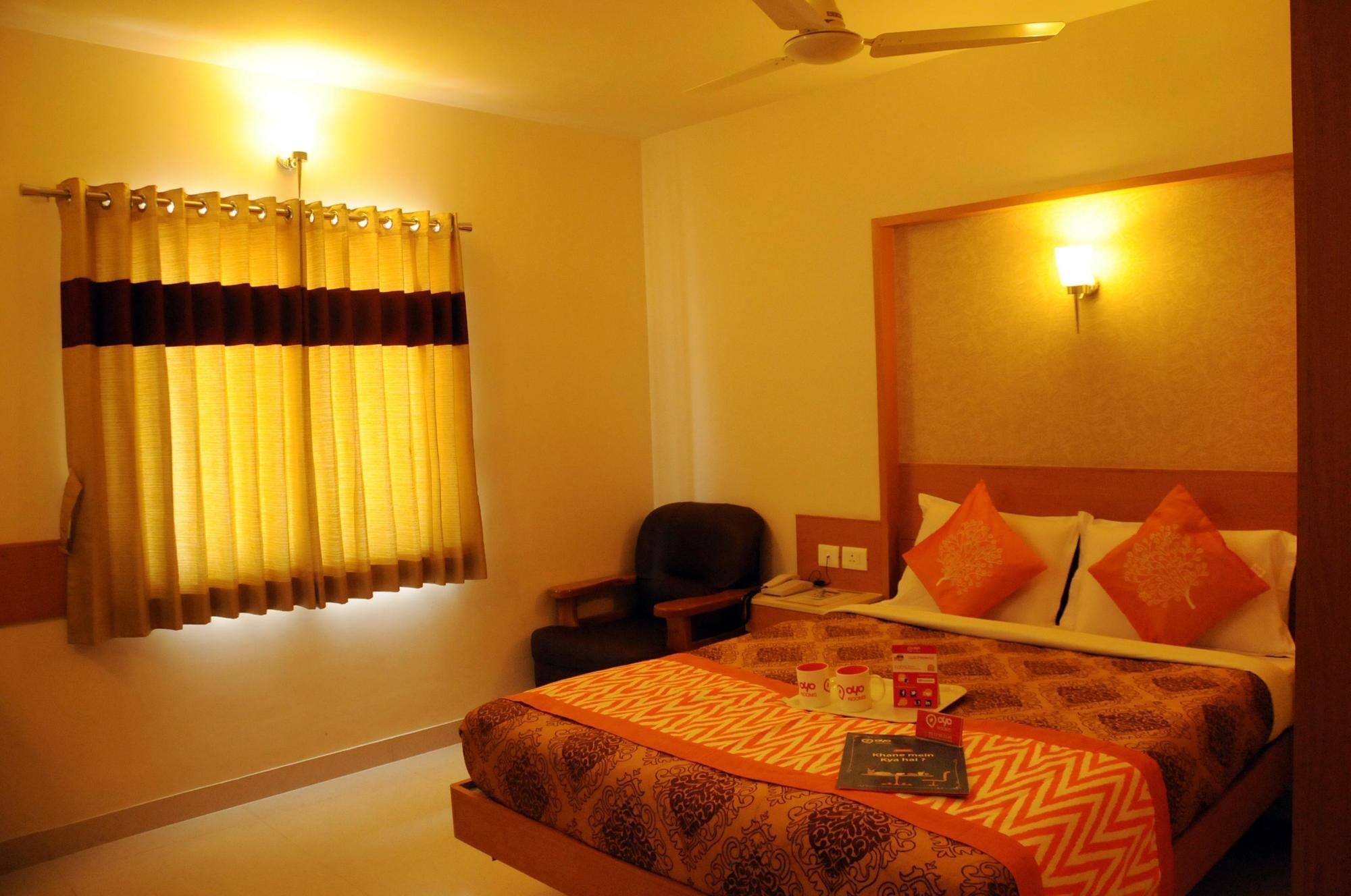 A hotel room with a bed, a chair, a desk with a phone, and a wall-mounted light.