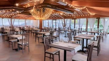 Un'ampia sala da pranzo con diversi tavoli e sedie, grandi finestre e un lampadario decorativo.