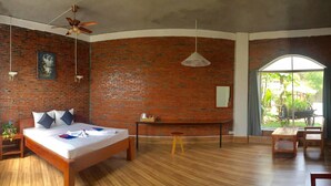 Una habitación con pared de ladrillo, una cama con almohadas, un ventilador de techo, piso de madera, una mesita con una planta y vistas a la vegetación exterior.