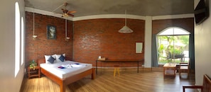 Una habitación con pared de ladrillo, una cama con almohadas, un ventilador de techo, piso de madera, una mesita con una planta y vistas a la vegetación exterior.