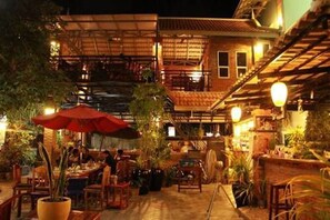 Un espacio de comedor al aire libre con mesas y sillas, rodeado de plantas en macetas y un paraguas rojo.