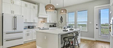 A modern kitchen with white cabinetry, a central island, pendant lighting, and a view through the window.