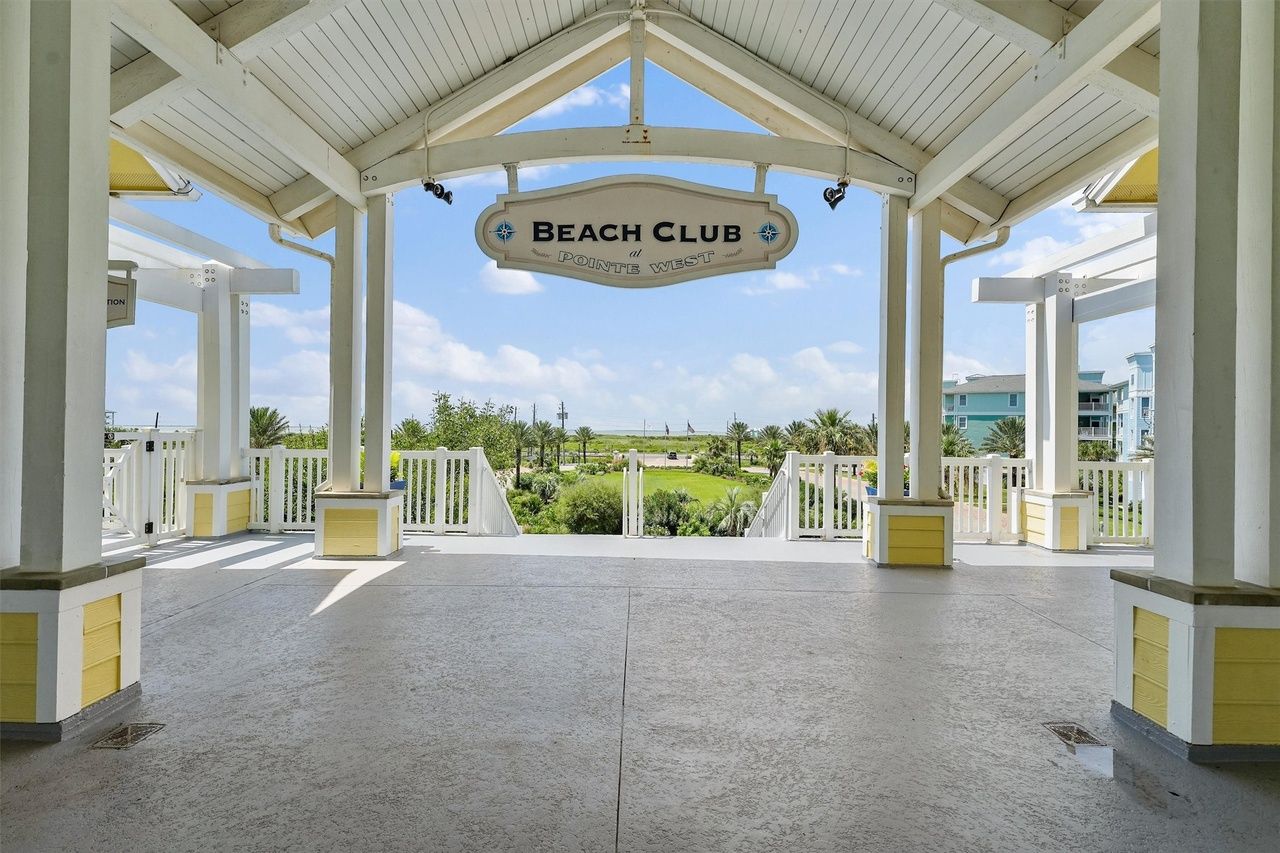 A covered outdoor area with a sign reading 'Beach Club at Pointe West,' featuring a view of a landscaped area and buildings in the background.
