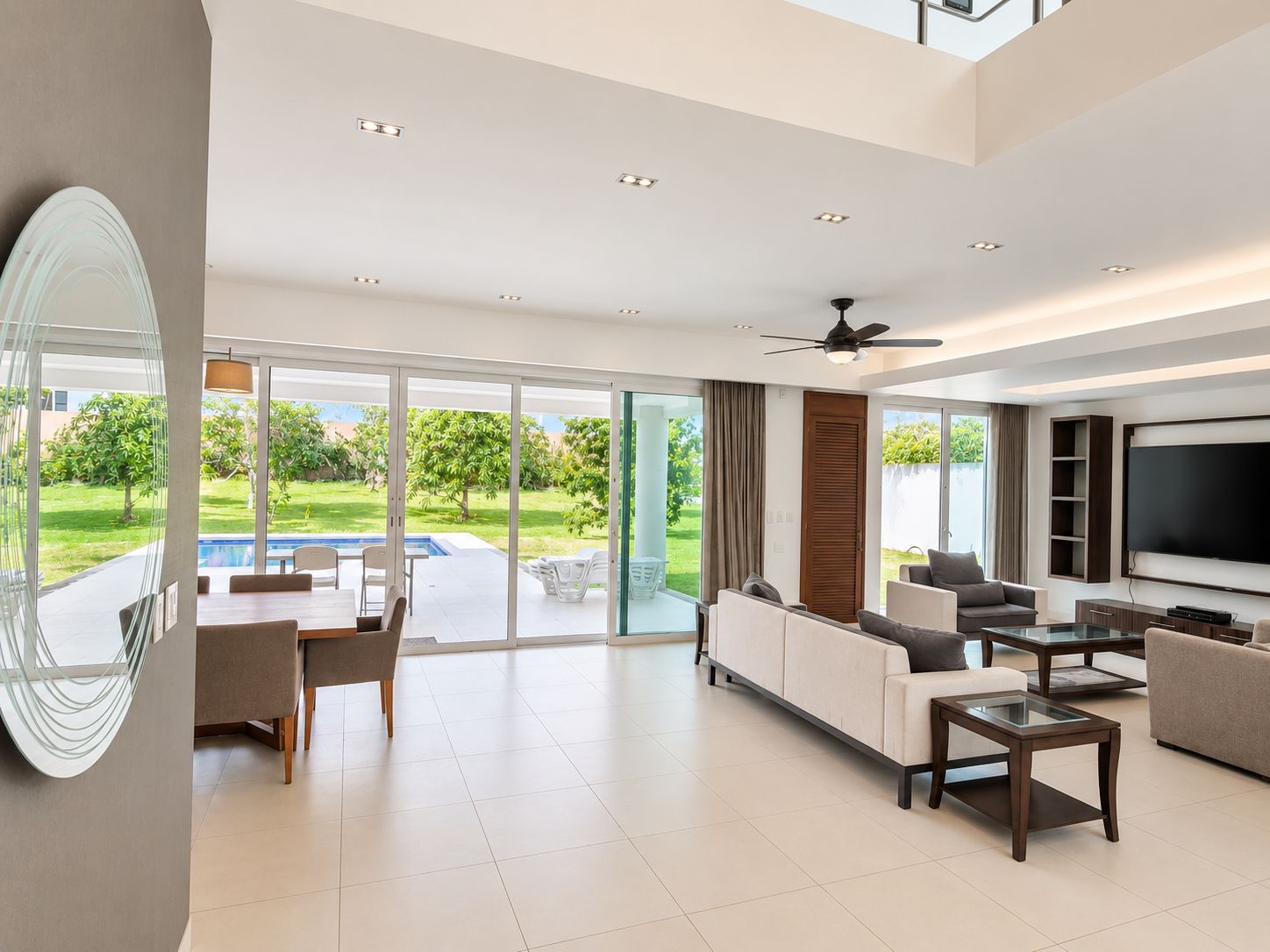 A modern living room with a flat-screen TV, comfortable seating, and a glass door leading to a pool area.