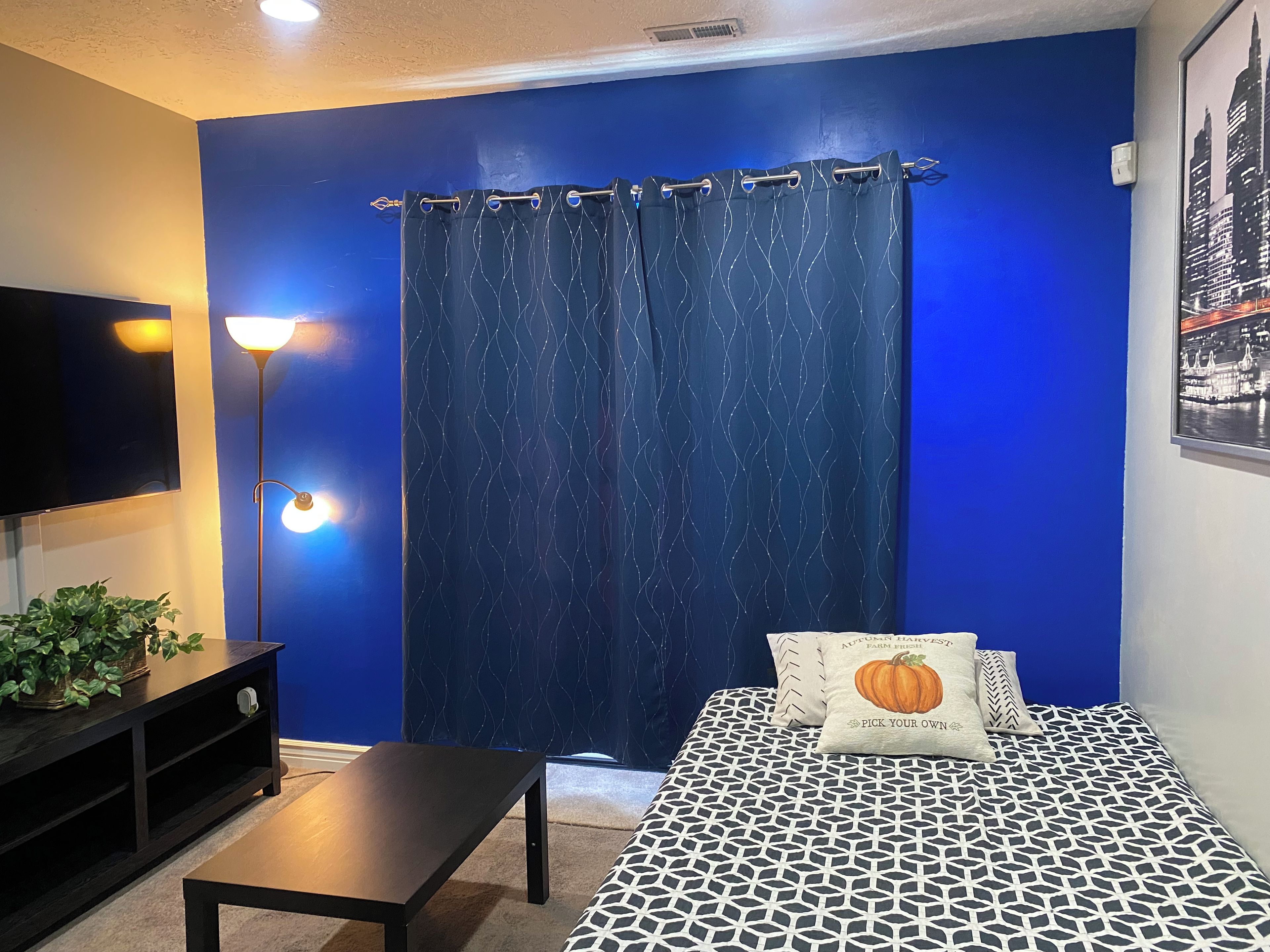 A room with a blue wall, a bed with patterned bedding, a black table, a floor lamp, and a television.