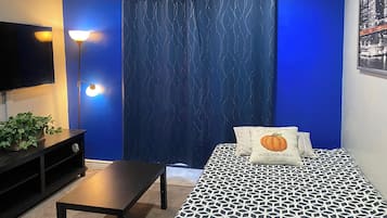A room with a blue wall, a bed with patterned bedding, a black table, a floor lamp, and a television.