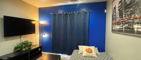 Une chambre avec un lit, une télévision, une plante et un tableau représentant un paysage urbain.