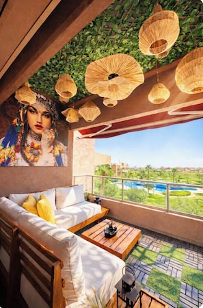A balcony with a sofa, hanging wicker lamps, and a view of a pool and greenery. - Beautiful apartment in downtown Marrakesh—3 outdoor pools, a golf course, and a terrace (Marrakech)