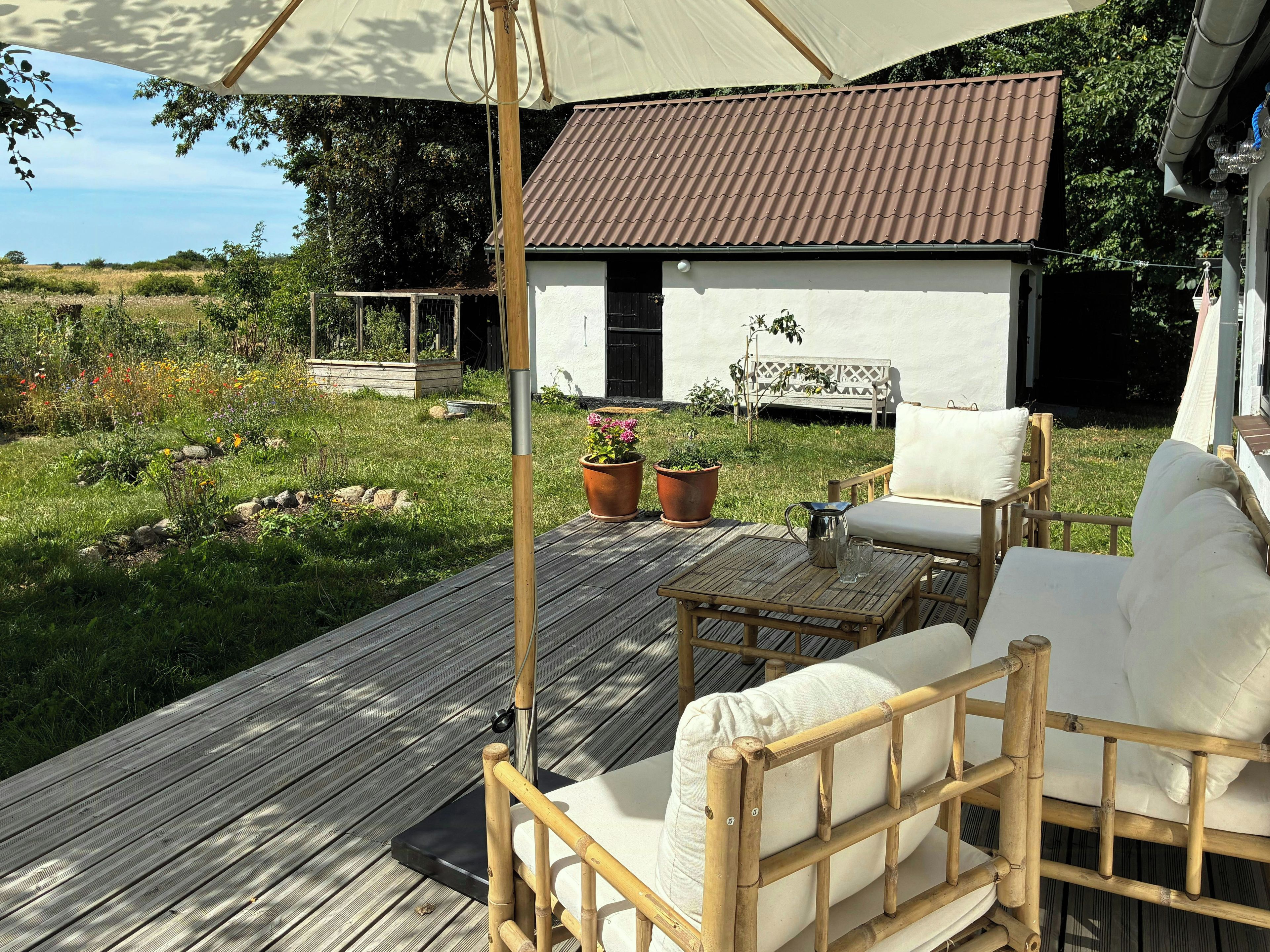 Una terraza de madera con mobiliario exterior, un edificio blanco con techo marrón y un jardín con diversas plantas.