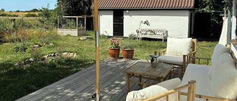 A wooden deck with outdoor furniture, a white building with a brown roof, and a garden with various plants.
