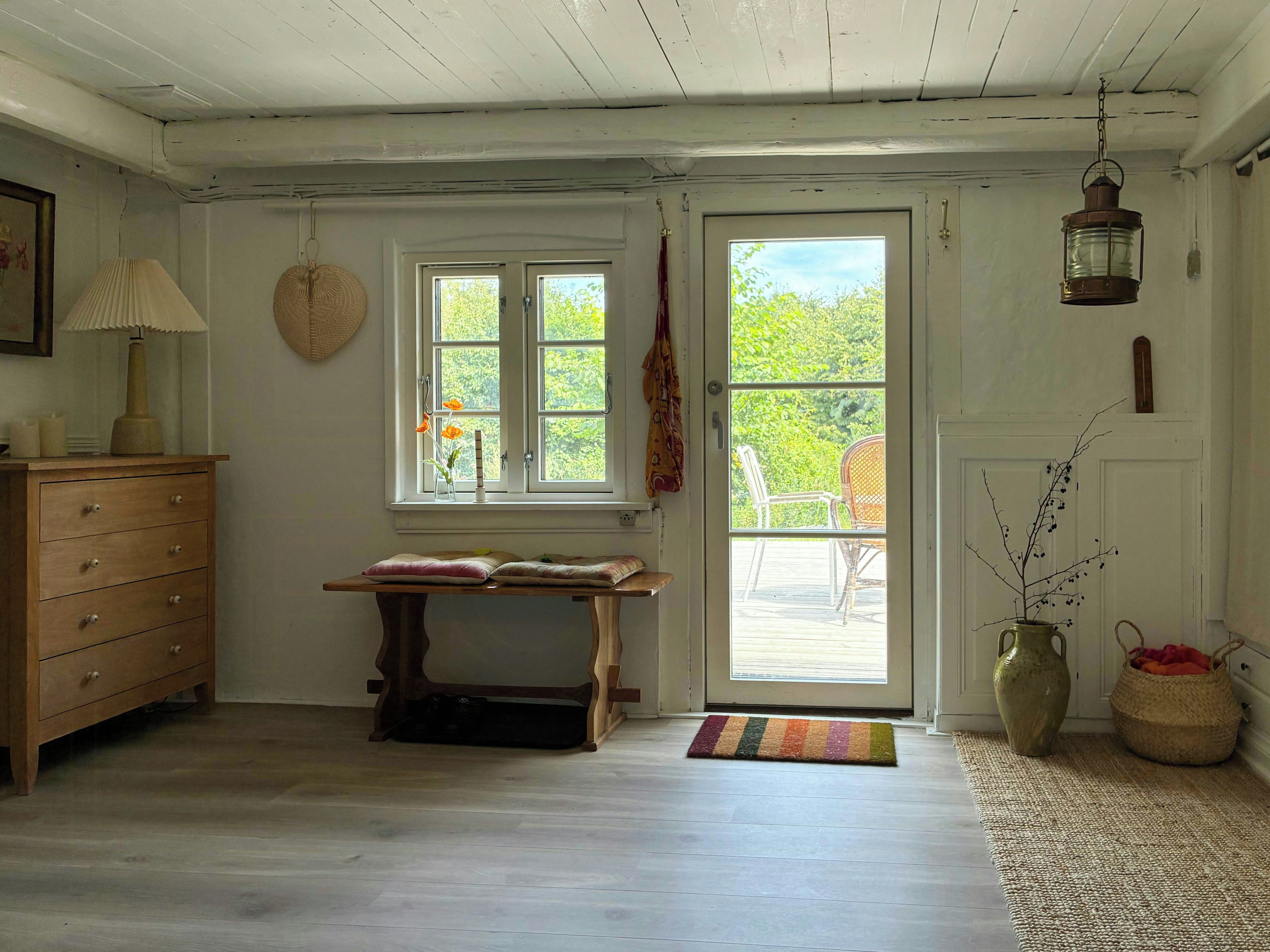 A room with a wooden dresser, a table with a rug, a window with a view, and a door leading outside.