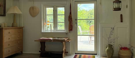 A room with a wooden dresser, a table with a rug, a window with a view, and a door leading outside.