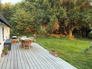 Une terrasse en bois avec une table et des chaises, entourée d’un jardin luxuriant et d’arbres.