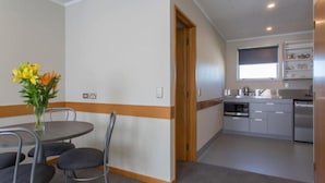 A room with a dining table, chairs, a vase of flowers, a kitchen with appliances, and a window. - Asure Dunedin Academy Court Motels (Dunedin)