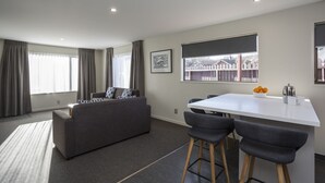 A modern living room with a sofa, a dining table with chairs, and a window with a view of a fence. - Asure Dunedin Academy Court Motels (Dunedin)