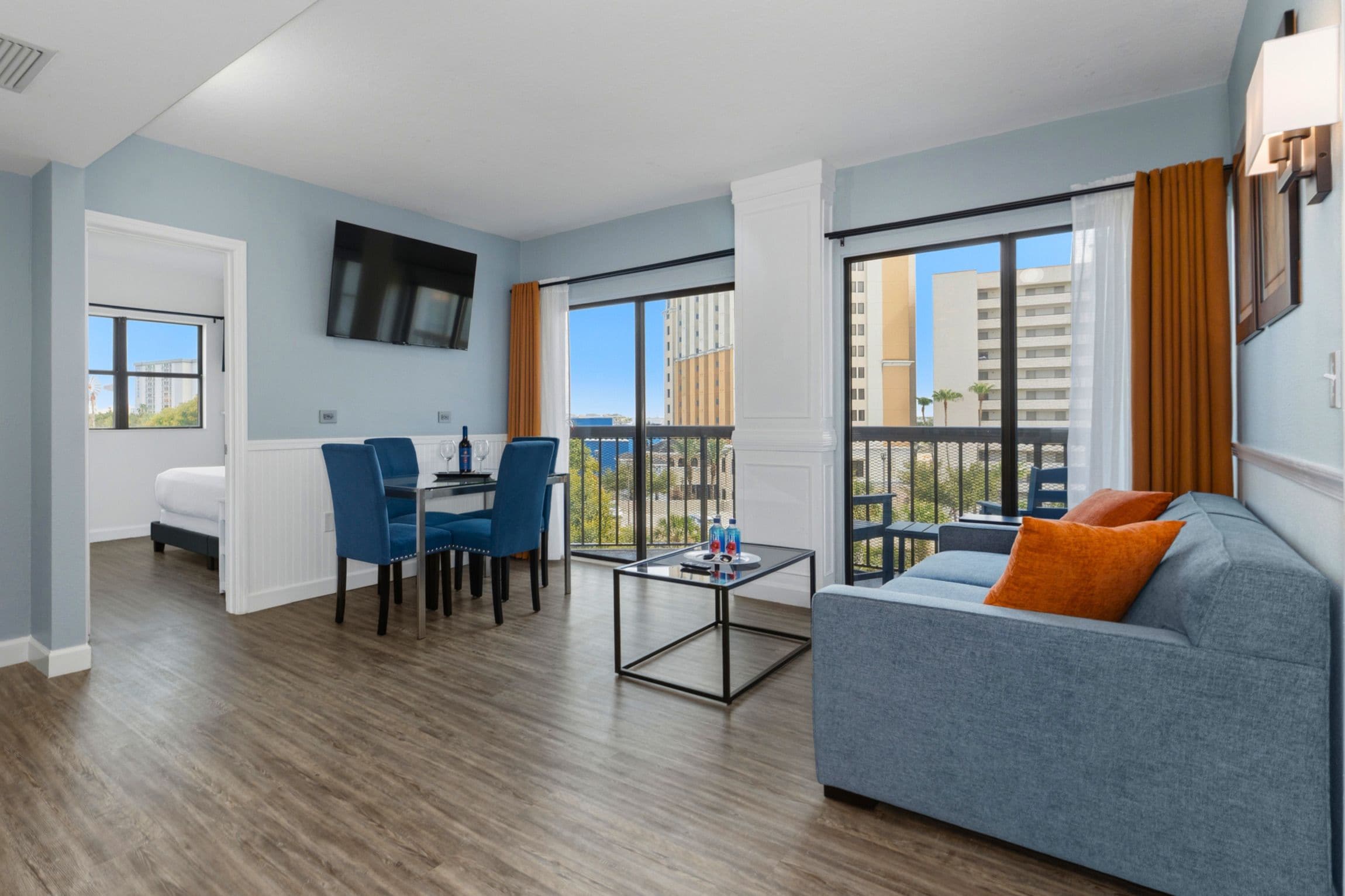 A modern living room with a sofa, a dining table with chairs, a TV mounted on the wall, and large windows offering a view of the city.
