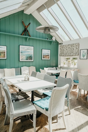 A dining area with white tables and wicker chairs, a green wall with framed artwork, and a ceiling fan. - The Cottage (Derby)