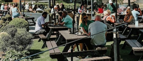 An outdoor patio area with multiple tables and umbrellas, surrounded by greenery and a clear sky.