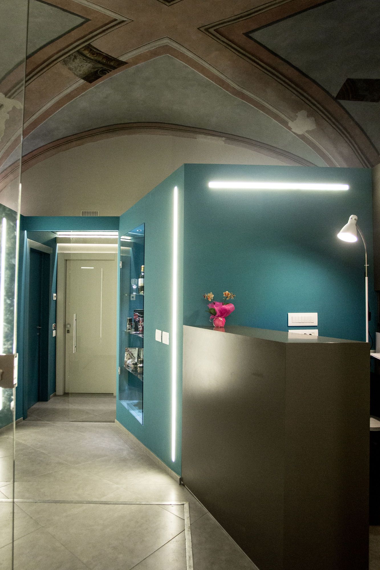 Un'area reception con una parete color petrolio, una scrivania e una lampada bianca, più un vaso con fiori.