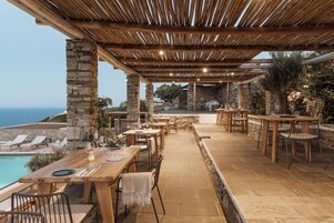 An outdoor dining area with stone pillars, wooden tables, and chairs, overlooking a pool and the sea.