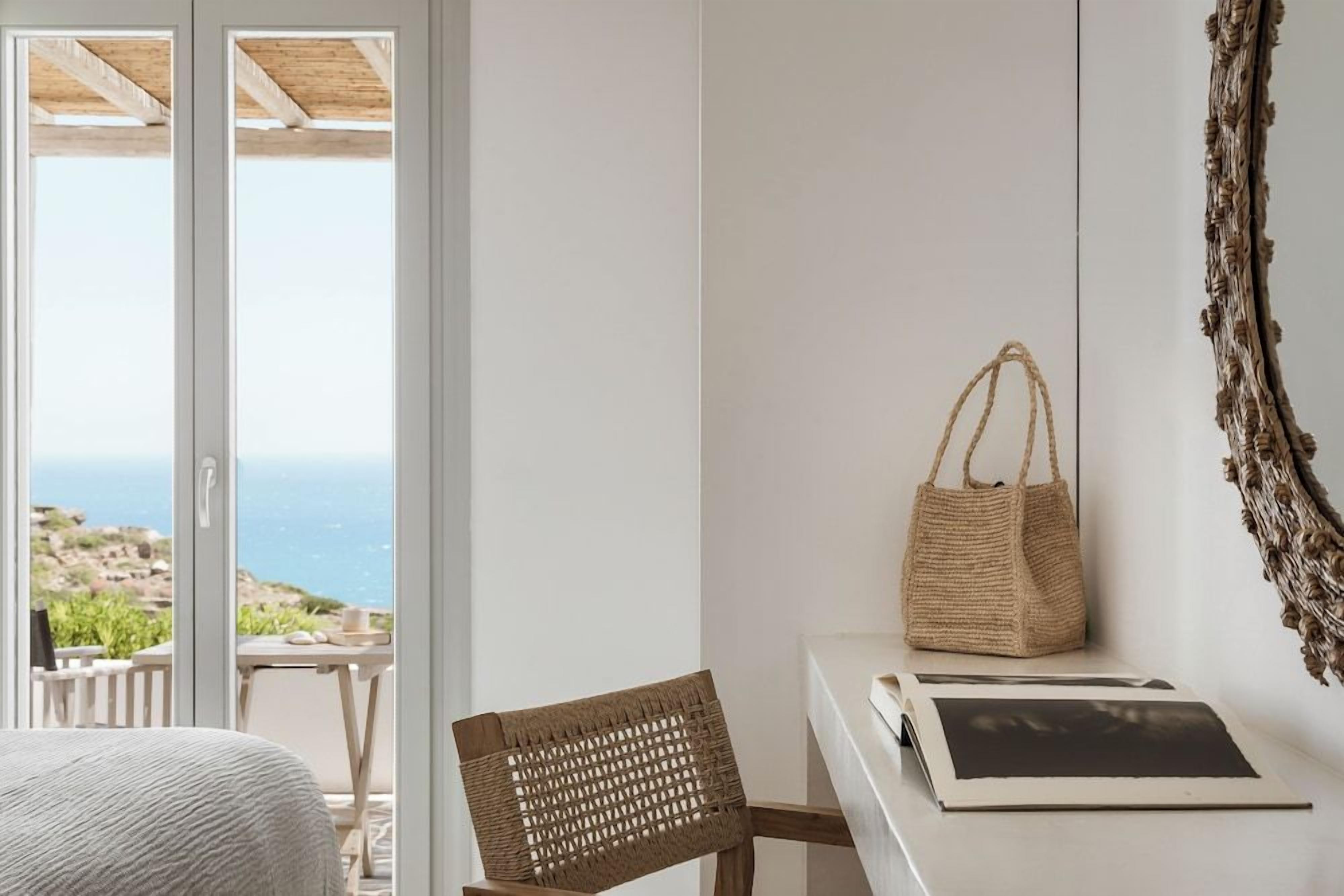 A room with a view of the sea, a wicker chair, a white table with a photo frame, and a woven bag.