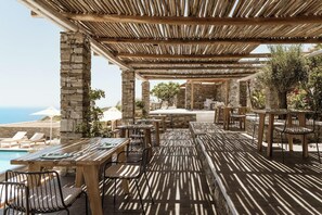 An outdoor dining area with stone pillars, wooden tables, and chairs, overlooking a pool and the sea. - Gerofinikas Complex (Sifnos)