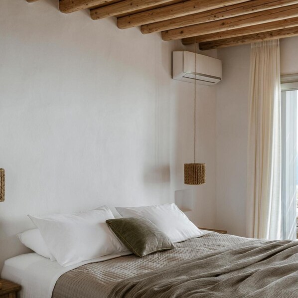 A bedroom with a bed, white pillows, a grey blanket, a hanging light fixture, and a window with curtains. - Gerofinikas Complex (Sifnos)