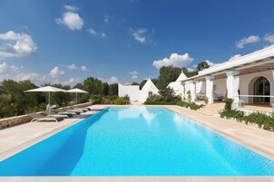 Una piscina con sillas de descanso y sombrillas, rodeada de edificios blancos y follaje.