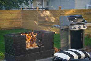 A barbecue grill with a built-in fire pit, a cushioned outdoor seating area, and a wooden fence in the background. - Serene 4-bedroom house with central AC and heating in charming Norwalk (Norwalk)