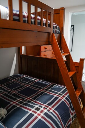 A bunk bed with a wooden ladder, a plaid-patterned bedspread, and a small wooden dresser.