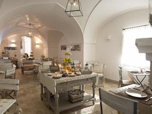 A dining area with tables set for meals, a chandelier, and a view of the kitchen in the background. - Masseria Don Luigi (Fasano)